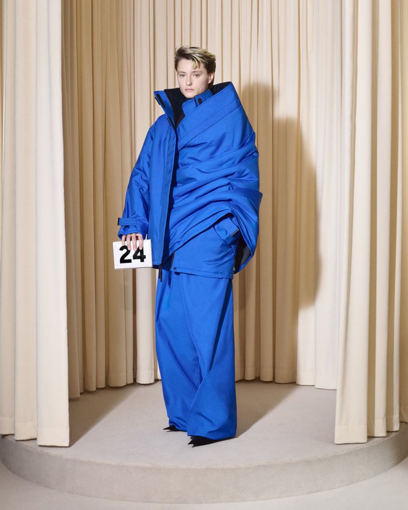 Fashion portrait of a person in an oversized blue puffer jacket and matching pants, holding a number card, standing in a studio.