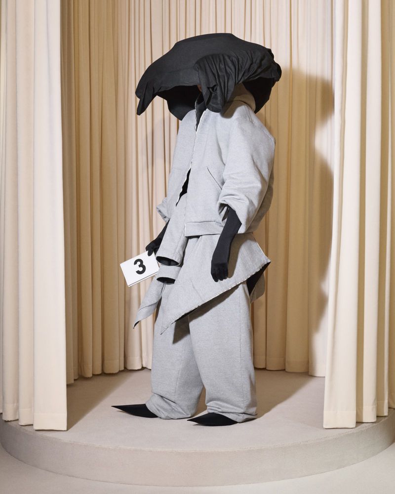 Person in oversized light grey suit with black gloves and hat-like head covering, standing on a platform with cream colored curtains.