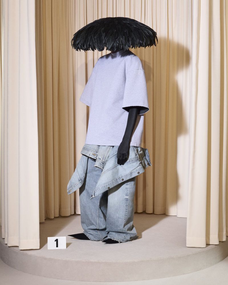 Fashion shot of model in oversized blue pants and top with feathered headpiece on a carpeted platform in front of a curtain.