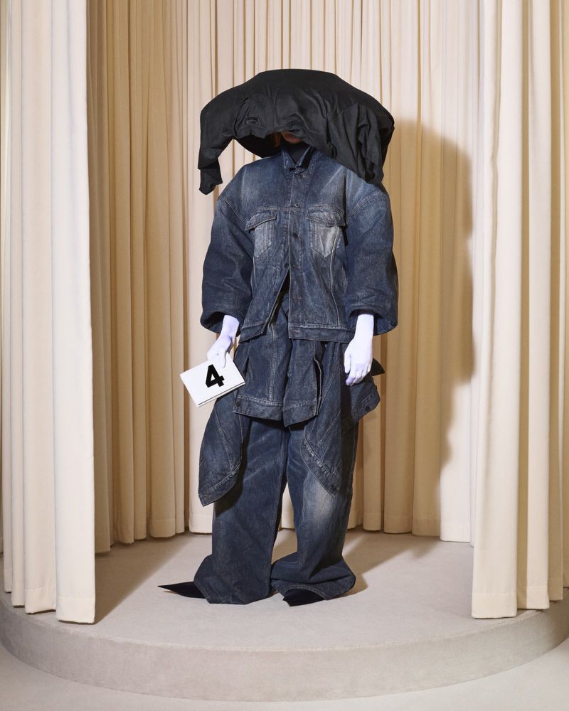 Person stands in denim outfit holding a white card with '4' on it, accessorized with a black jacket on their head, and against a neutral curtain.