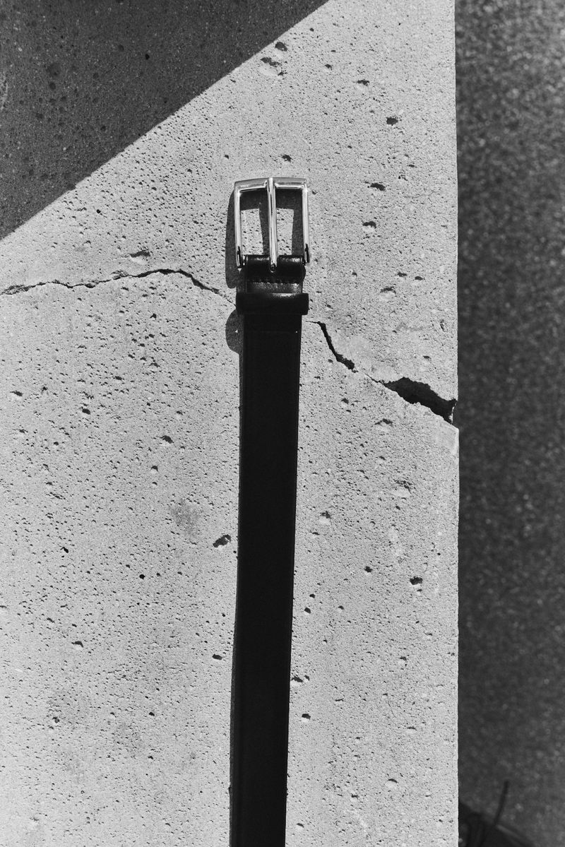 Close-up shot of a black belt with a silver buckle leaning against a textured, cracked concrete wall in black and white.