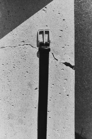 Close-up shot of a black belt with a silver buckle leaning against a textured, cracked concrete wall in black and white.