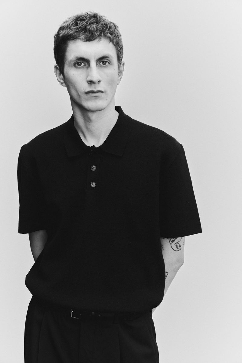 Portrait of an adult male in a black knit polo shirt and dark trousers, posing in a studio with a light gray seamless background.