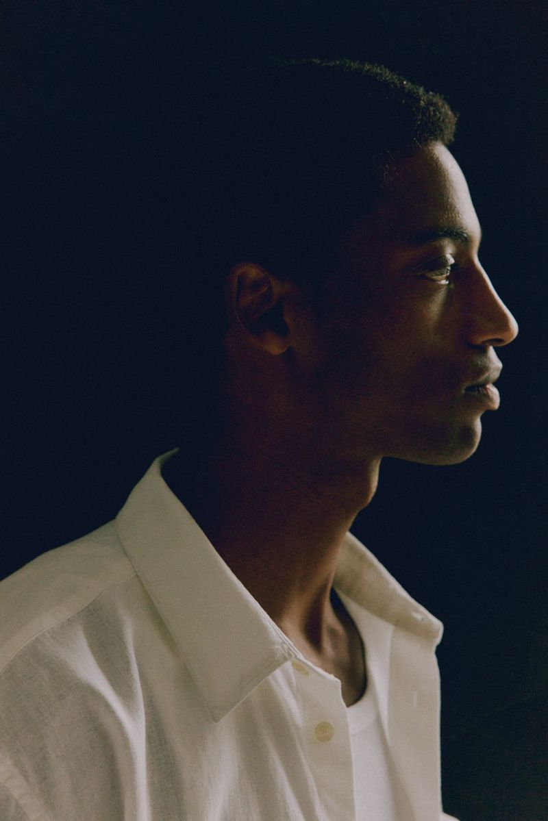 Portrait of a young adult Black male in profile, wearing a white collared shirt, set against a dark background with soft, directional lighting.