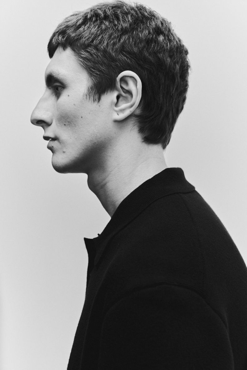 Monochrome portrait of an adult male in profile, wearing a dark collared shirt, set against a neutral grey backdrop.