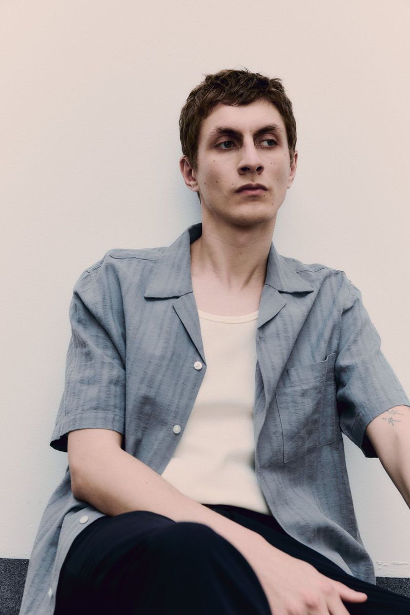 Portrait of a young adult man in a collared short-sleeved shirt and t-shirt, casually sitting indoors, looking off to the side.