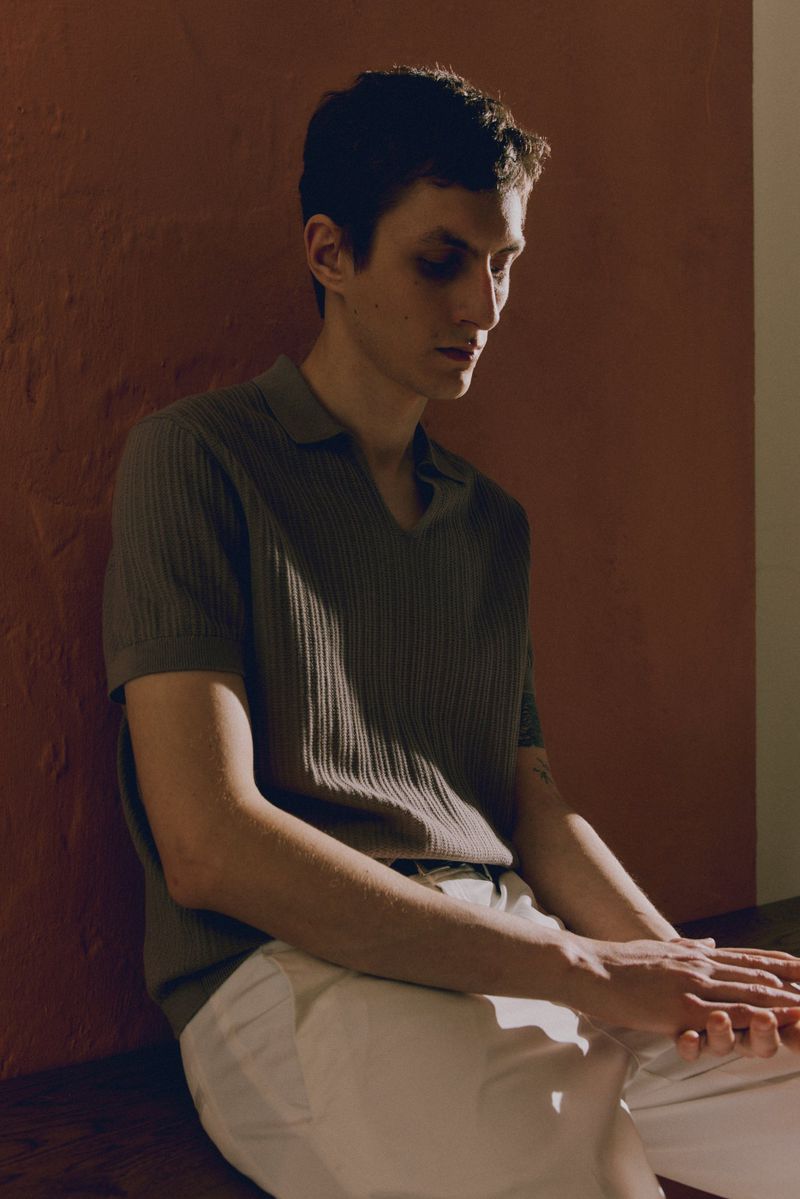 Indoor portrait of a light-skinned adult male in a ribbed polo shirt and cream pants, seated against a warm-toned wall, looking down.