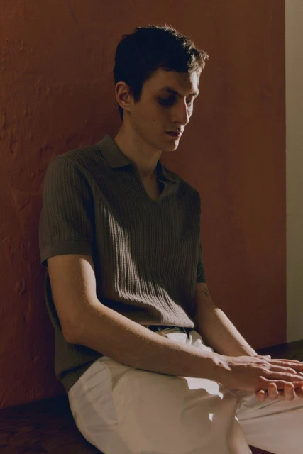 Indoor portrait of a light-skinned adult male in a ribbed polo shirt and cream pants, seated against a warm-toned wall, looking down.