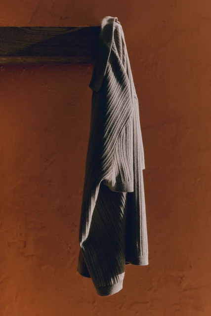 Knitted lounge wear set hangs on wooden bar against earth-toned wall with shadows and textures adding depth and warmth to the apparel.