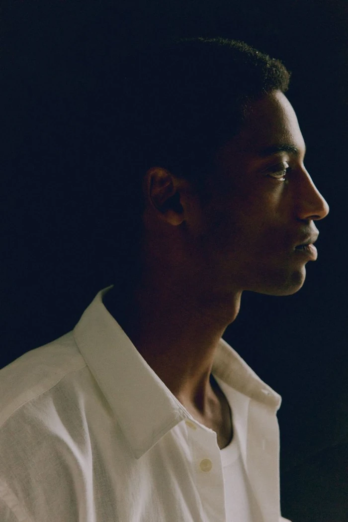 Profile portrait of a young man wearing a white collared shirt against a dark background for the H&M Man Spring 2025 lookbook.