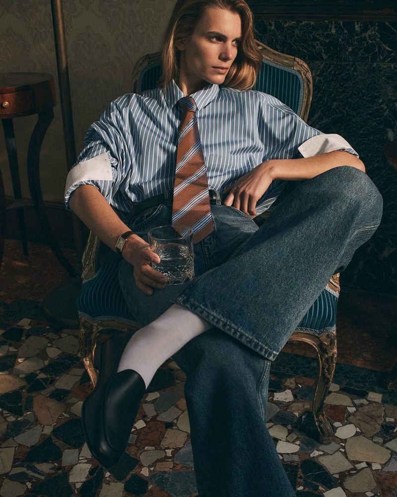 Stylish portrait of a blonde-haired person in a striped shirt and jeans, seated on a blue chair, holding a glass of water.