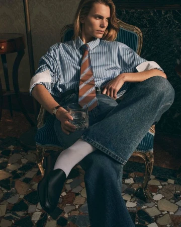 Stylish portrait of a blonde-haired person in a striped shirt and jeans, seated on a blue chair, holding a glass of water.