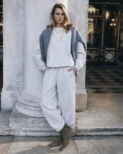 Woman in casual white sweatsuit with a grey sweater draped over her shoulders standing near a pillar in an architectural setting.