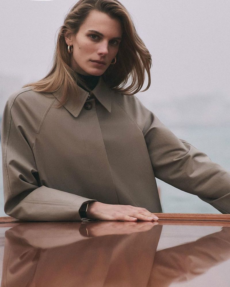 Adult woman poses in a neutral-colored coat outdoors, with water in the background on a cloudy day, wearing gold earrings.
