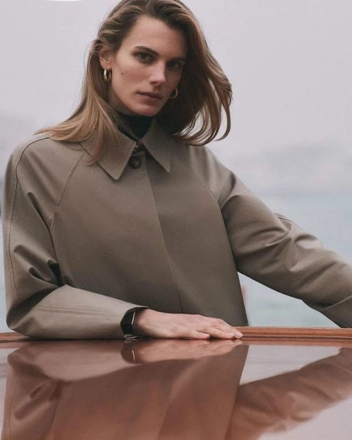 Adult woman poses in a neutral-colored coat outdoors, with water in the background on a cloudy day, wearing gold earrings.