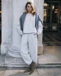 Model leaning against a stone column wearing an off-white matching sweatshirt and sweatpants with layered pearl necklaces.