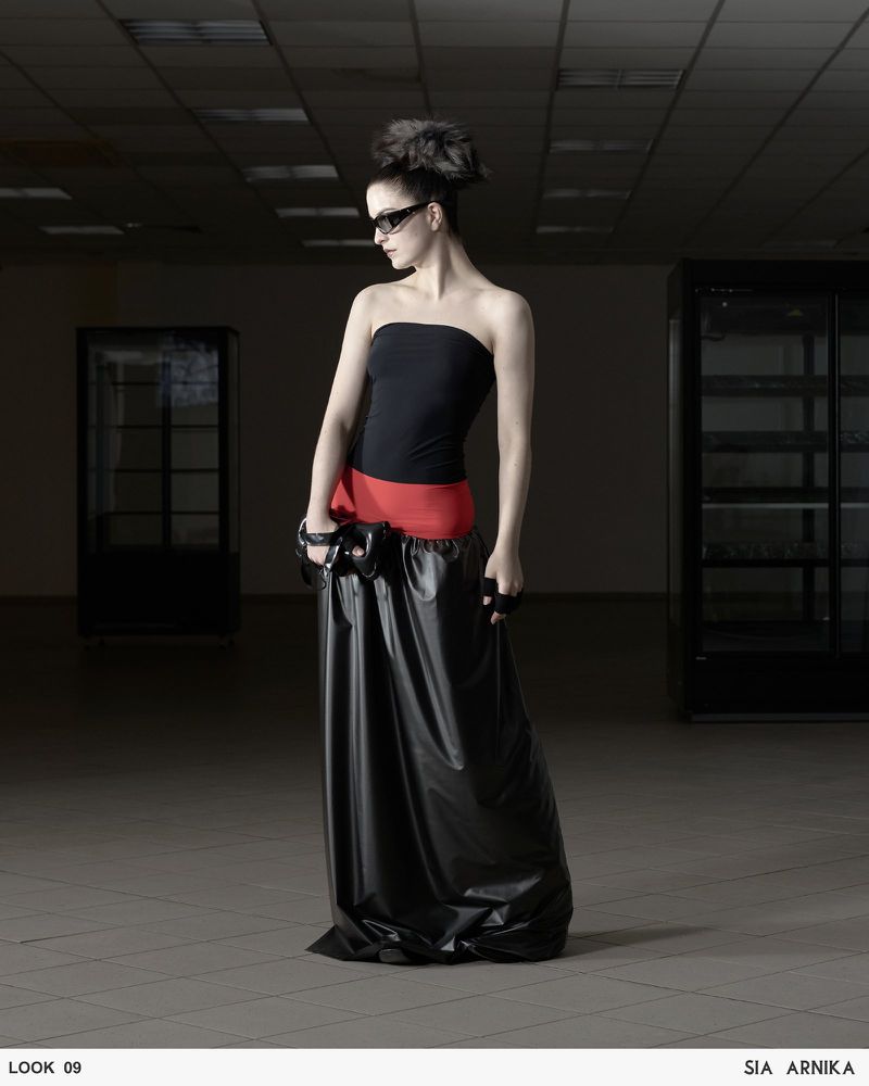A woman poses in a black strapless top and a voluminous black skirt with a red waistband against a dimly lit indoor backdrop.