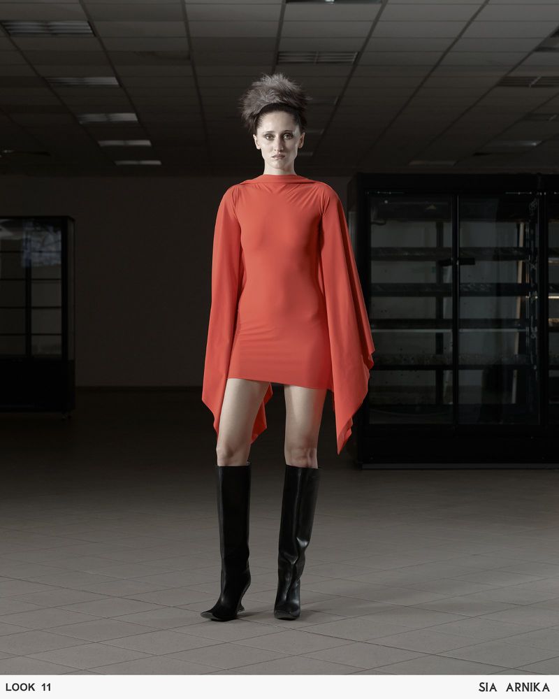 Adult female fashion model in a short orange dress and black knee-high boots stands in an empty interior space.
