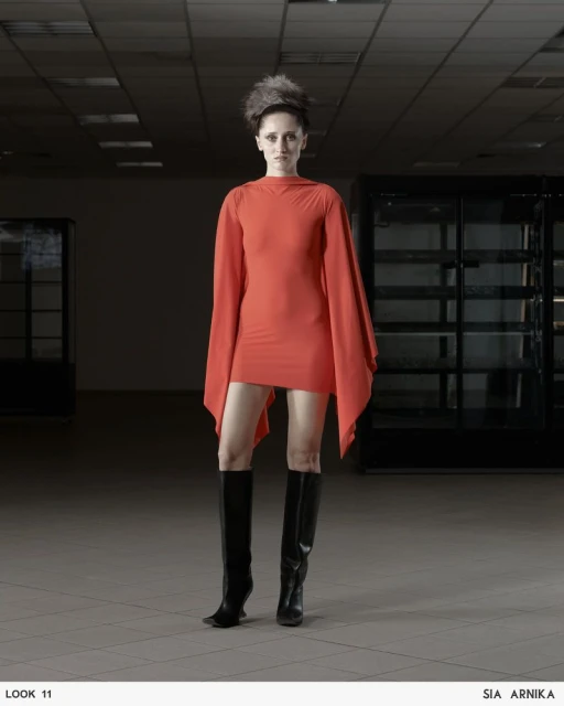 Adult female fashion model in a short orange dress and black knee-high boots stands in an empty interior space.