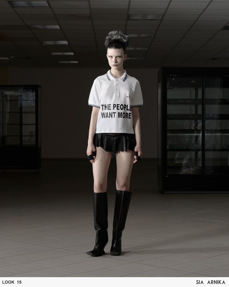 Fashion shot of a slender adult female wearing a white polo shirt with black trim, a black skirt, and black boots in an empty space.