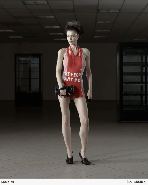 A fashion shot of a woman in a short, red and white dress standing in a mostly empty room, holding a black purse in front of her.
