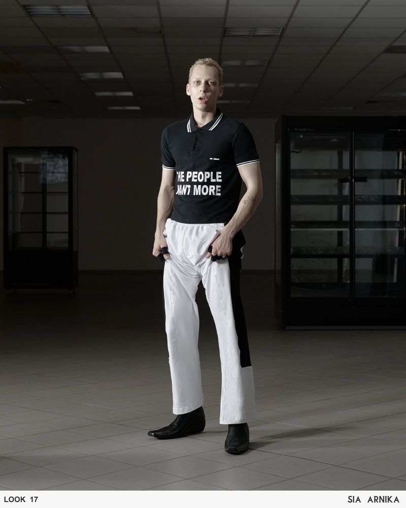 Full-length shot of a fair-skinned adult male in a black polo shirt with the text 'THE PEOPLE WANT MORE,' and white pants, posing indoors.