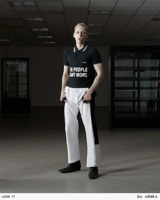 Full-length shot of a fair-skinned adult male in a black polo shirt with the text 'THE PEOPLE WANT MORE,' and white pants, posing indoors.