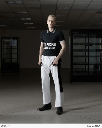 Full-length shot of a fair-skinned adult male in a black polo shirt with the text 'THE PEOPLE WANT MORE,' and white pants, posing indoors.