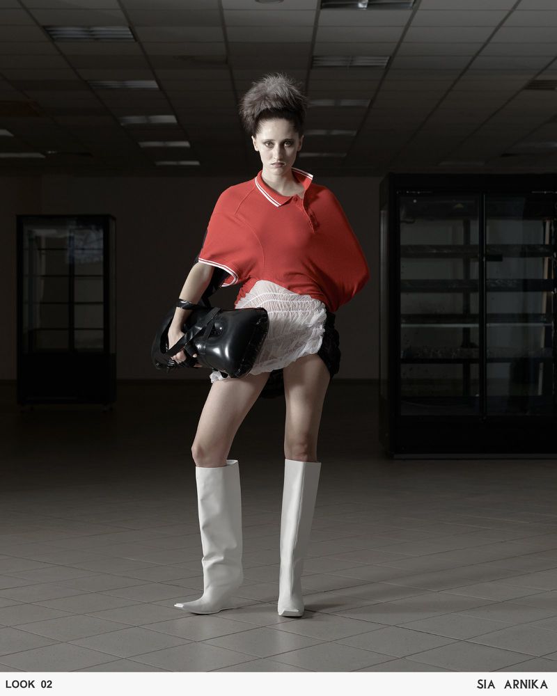 Fashionable woman with a retro hairstyle, white boots, red shirt, and black bag poses in a sparsely decorated room.