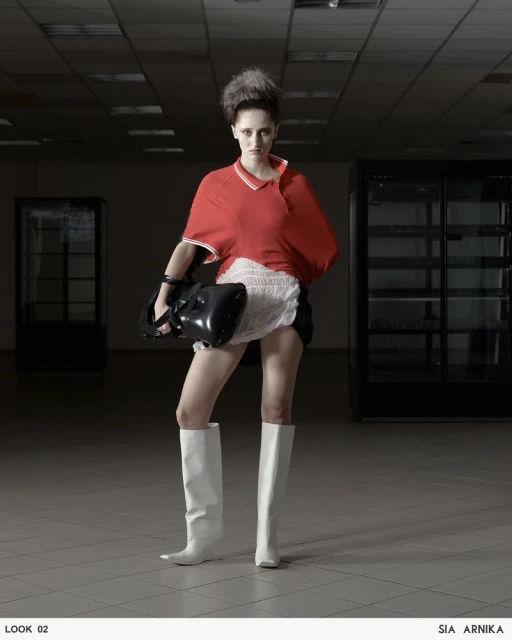 Fashionable woman with a retro hairstyle, white boots, red shirt, and black bag poses in a sparsely decorated room.