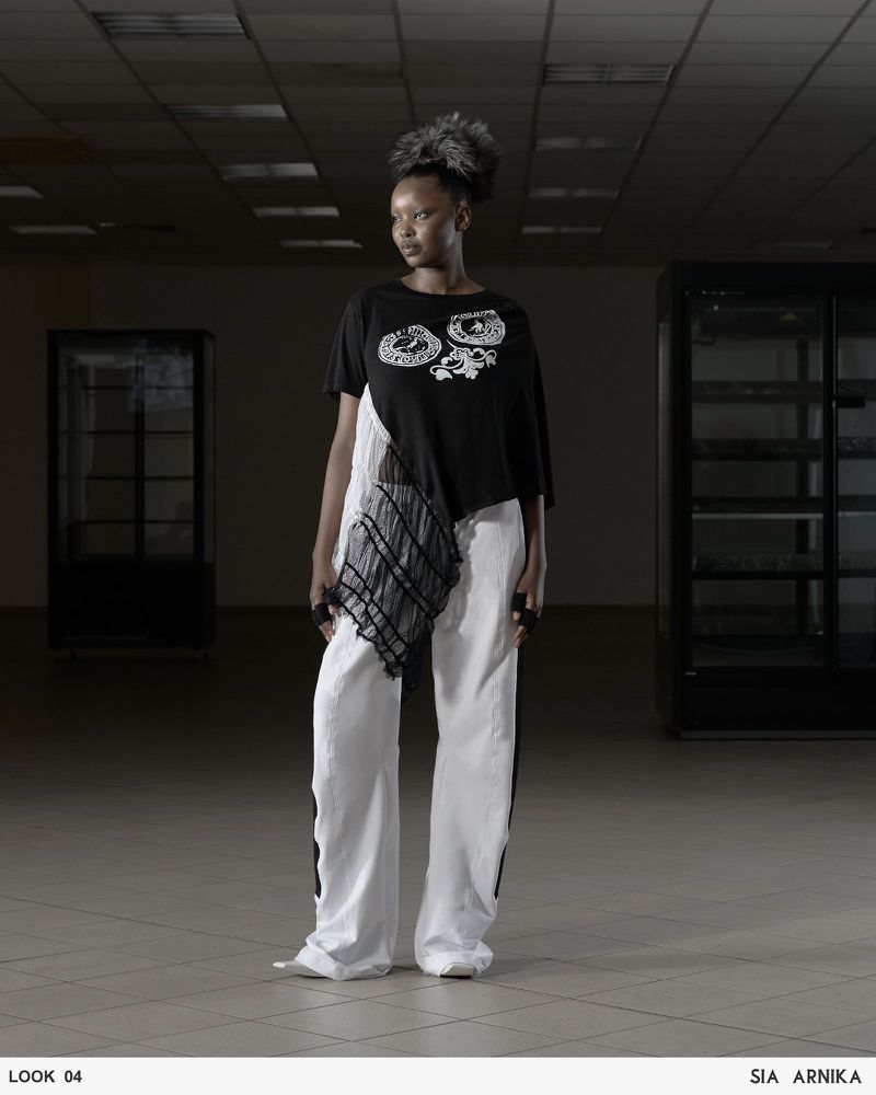 A stylish Black woman poses indoors wearing an asymmetrical black top, white pants, and unique hair, standing against a neutral backdrop.