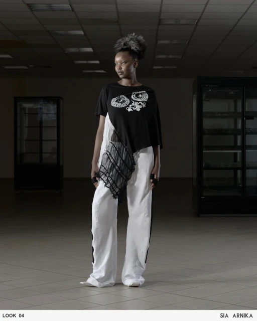 A stylish Black woman poses indoors wearing an asymmetrical black top, white pants, and unique hair, standing against a neutral backdrop.