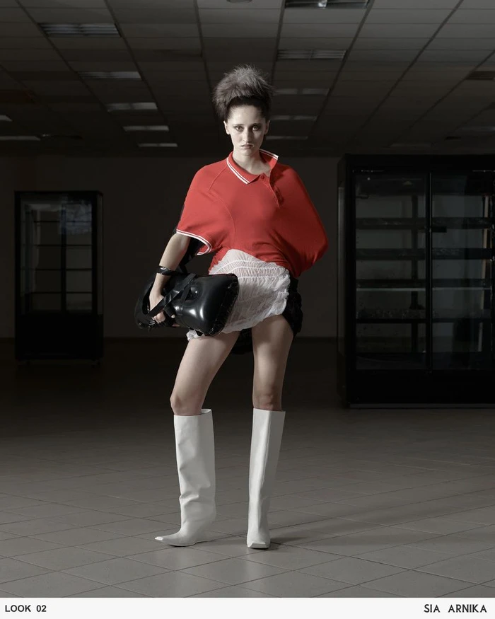 Model wearing a red polo top, white ruffled skirt, and white boots holding a black accessory inside a sparse room.