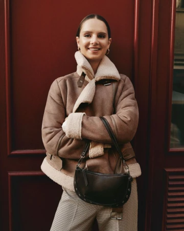 Adult woman posing in a stylish shearling jacket with a black purse against a red door background, fashion advertisement