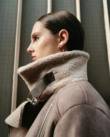 Profile portrait of a woman wearing a taupe shearling collar jacket and gold hoop earrings against a slatted architectural background.