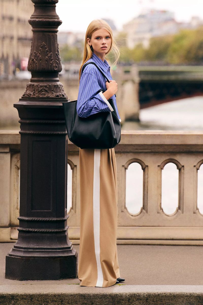 Outdoor fashion shot of a blonde woman in blue striped shirt and tan pants with a black tote bag, standing on a city bridge.