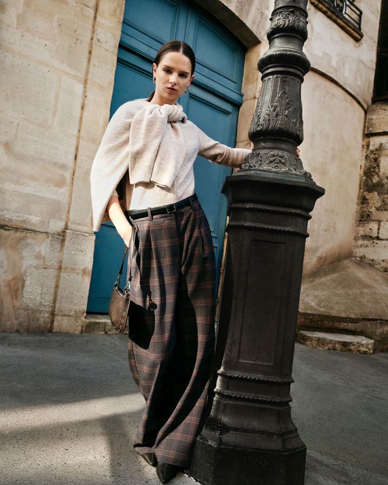 A woman poses in wide-leg, plaid pants and a sweater on a city street next to a black lamppost and a blue door, styled for fashion.