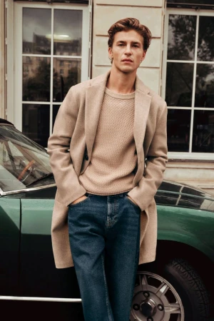 A male model wearing a beige overcoat and knitted sweater standing against a car in front of a building.