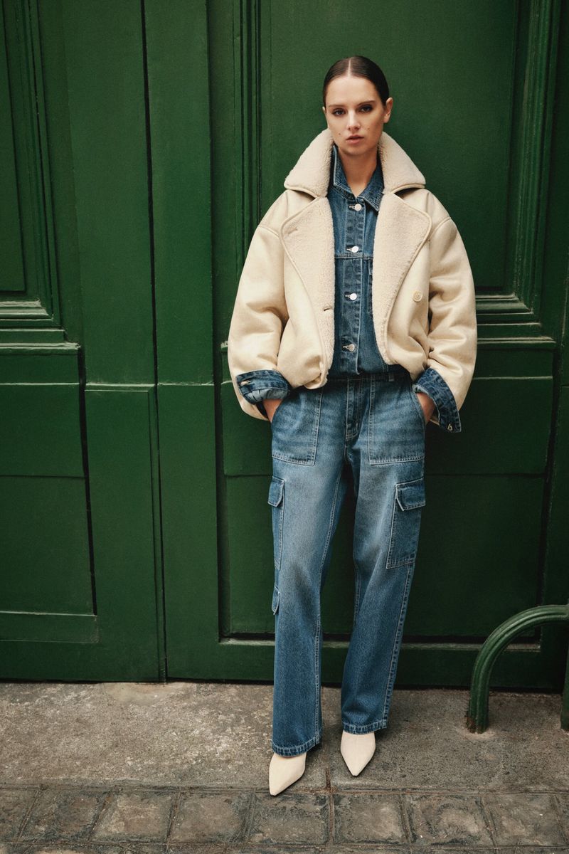 Woman in denim shirt and cargo jeans, wearing a cream shearling jacket, posing against a green door in an outdoor setting.