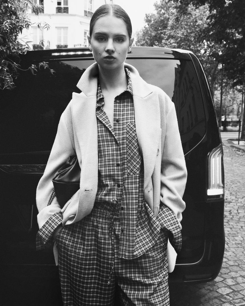 Black and white portrait of a woman in plaid shirt and coat, standing next to a black van in an urban environment on a cloudy day.