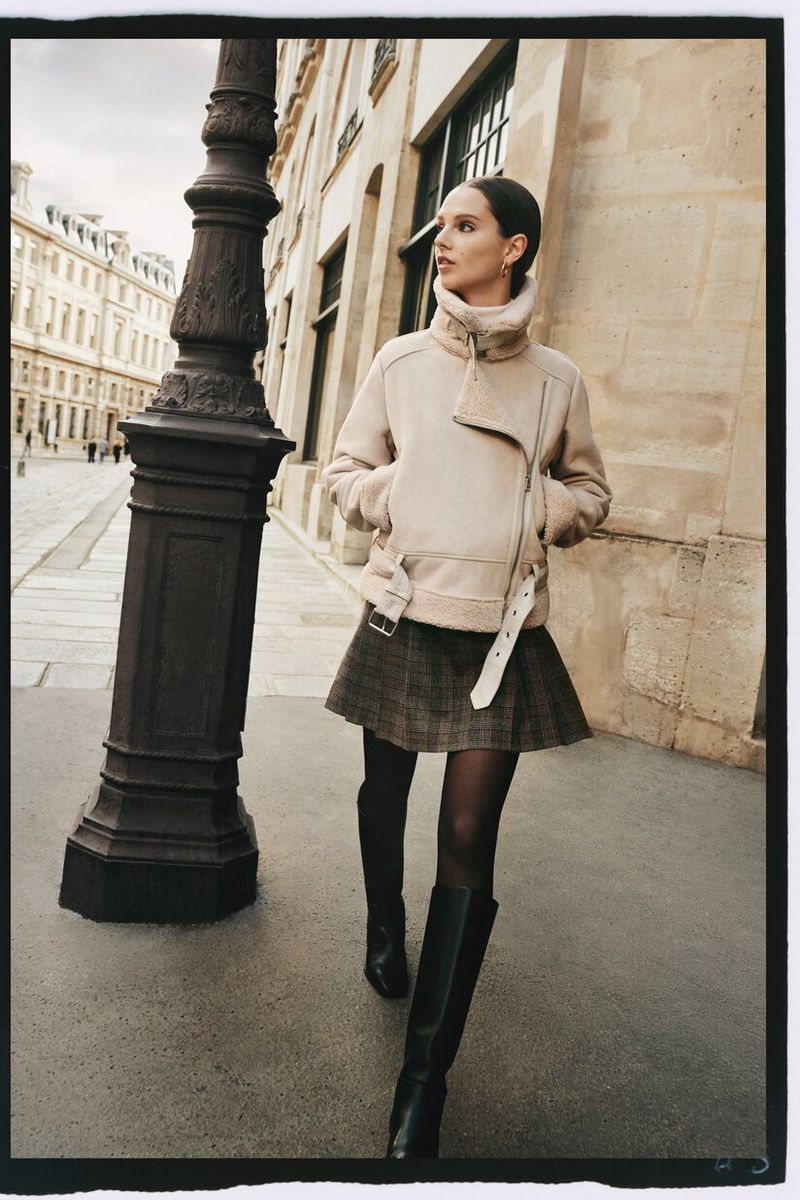 Woman in a beige jacket and plaid skirt walks on a European-style street next to a black lamp post on an overcast day.