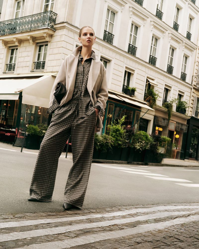 Fashionable woman in a plaid jumpsuit and cream jacket posing on a city street with European architecture in the background.