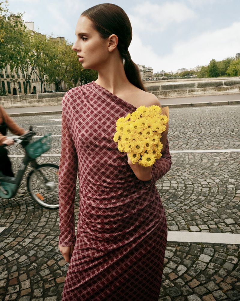 A woman stands on a cobblestone street in a red dress, holding yellow flowers, with a city scene and bicycle blurred in the background.