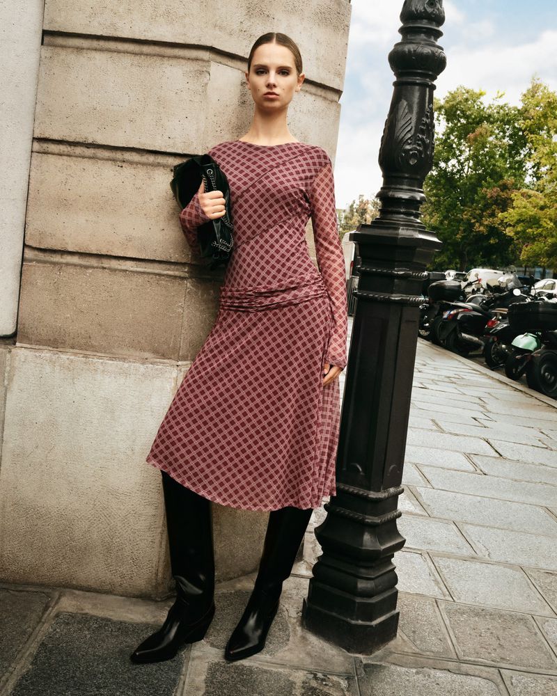 An adult female fashion model poses outdoors wearing a long-sleeved patterned dress and tall black boots, carrying a clutch purse.
