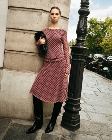 An adult female fashion model poses outdoors wearing a long-sleeved patterned dress and tall black boots, carrying a clutch purse.
