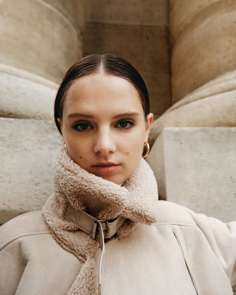 Close-up portrait of a young adult woman with neutral expression, wearing a sherpa-lined jacket against a beige wall with architectural columns.