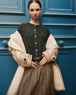 Model wearing a dark grey cardigan and shearling-lined coat standing before a blue double door
