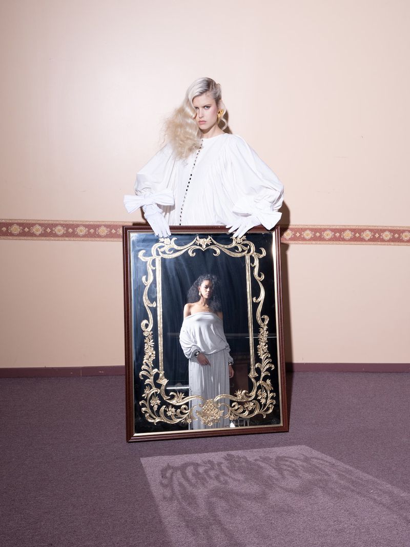 Woman poses with a large ornate mirror reflecting an image of another woman in a white dress against a peach-colored wall and purple carpet.
