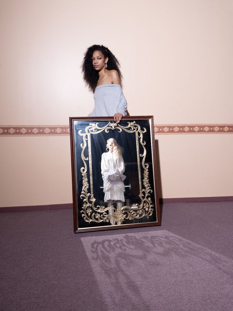 A young adult woman with dark curly hair wearing an off-the-shoulder top holding a framed mirror reflecting a woman in white clothing.