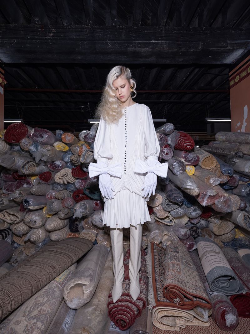 Fashion model in an ivory dress stands in front of rolled carpets in a dimly lit space, showcasing a blend of haute couture.